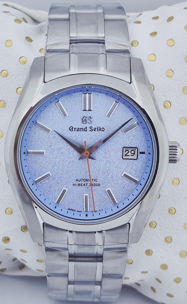 May include: A silver-toned Grand Seiko automatic wristwatch with a light blue dial. The watch features a date display, silver hands, and a silver bracelet. The dial has the text "Grand Seiko" and "Automatic Hi-Beat 36000".