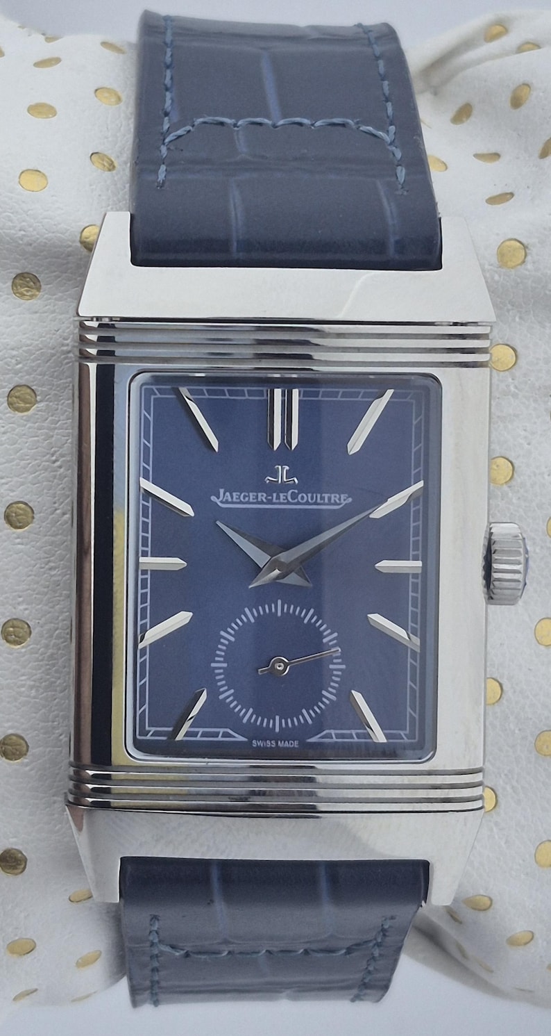 Pu&ograve; includere: Un orologio da polso rettangolare in acciaio inossidabile con quadrante blu navy e cinturino in pelle abbinato. Il quadrante dell'orologio presenta lancette e indici argentati, un piccolo quadrante dei secondi e il logo Jaeger-LeCoultre.
