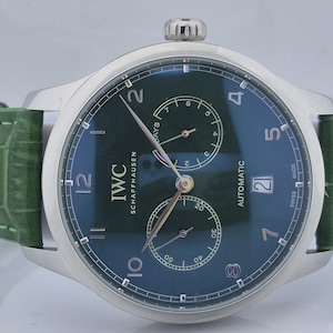 May include: A silver-cased IWC Schaffhausen automatic watch with a dark green dial and a green leather strap. The watch face features sub-dials, numerals, and the date. The strap is a textured green leather.