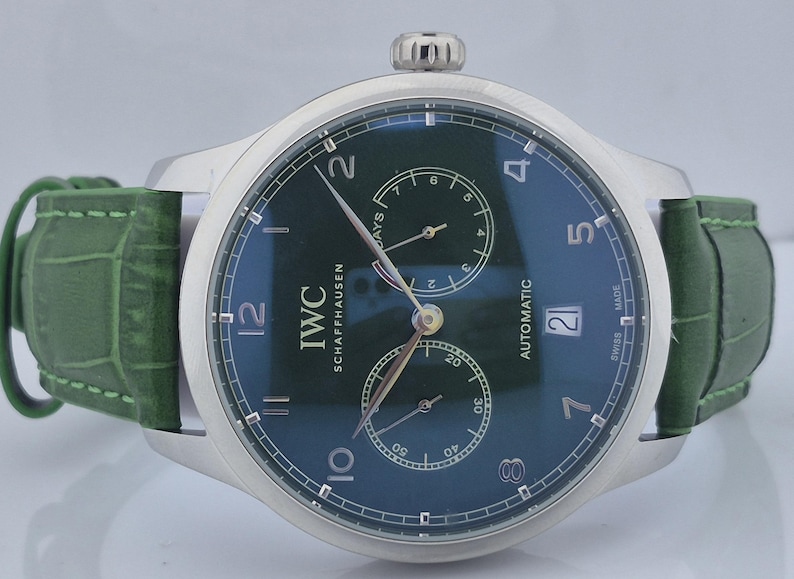 May include: A silver-cased IWC Schaffhausen automatic watch with a dark green dial and a green leather strap. The watch face features sub-dials, numerals, and the date. The strap is a textured green leather.