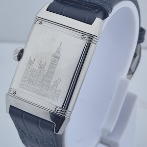 Pu&ograve; includere: Un orologio da polso rettangolare argentato con cinturino in pelle blu scuro. Il quadrante dell'orologio presenta un'immagine incisa di un edificio con un'alta torre dell'orologio. L'orologio &egrave; esposto su un supporto bianco.