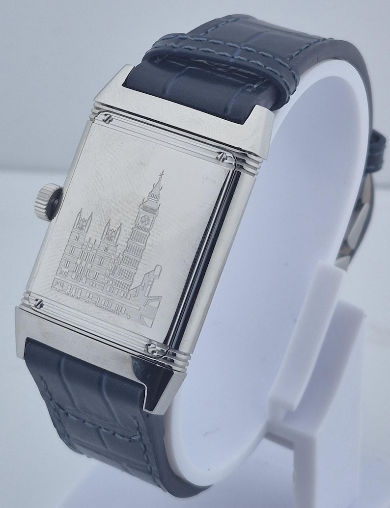 Pu&ograve; includere: Un orologio da polso rettangolare argentato con cinturino in pelle blu scuro. Il quadrante dell'orologio presenta un'immagine incisa di un edificio con un'alta torre dell'orologio. L'orologio &egrave; esposto su un supporto bianco.