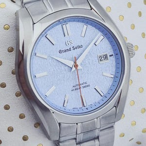 May include: A silver-toned Grand Seiko automatic wristwatch with a light blue dial. The watch features a date display, silver hands, and a silver bracelet. The dial has the text "Grand Seiko" and "AUTOMATIC HI-BEAT 36000".