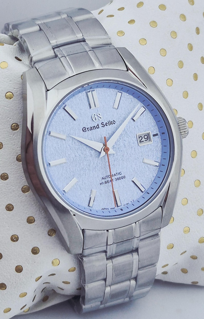 May include: A silver-toned Grand Seiko automatic wristwatch with a light blue dial. The watch features a date display, silver hands, and a silver bracelet. The dial has the text "Grand Seiko" and "AUTOMATIC HI-BEAT 36000".