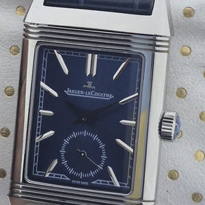 Pu&ograve; includere: Un orologio Jaeger-LeCoultre rettangolare in acciaio inossidabile con quadrante blu navy e cinturino in pelle abbinato. L'orologio presenta lancette e indici argentati, un piccolo quadrante dei secondi e la scritta "SWISS MADE".