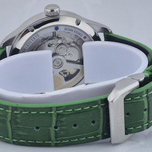 May include: A silver-toned wristwatch with a green leather band. The watch face is visible, showcasing the intricate internal mechanisms. The green band has a textured, crocodile-like pattern and a silver buckle.