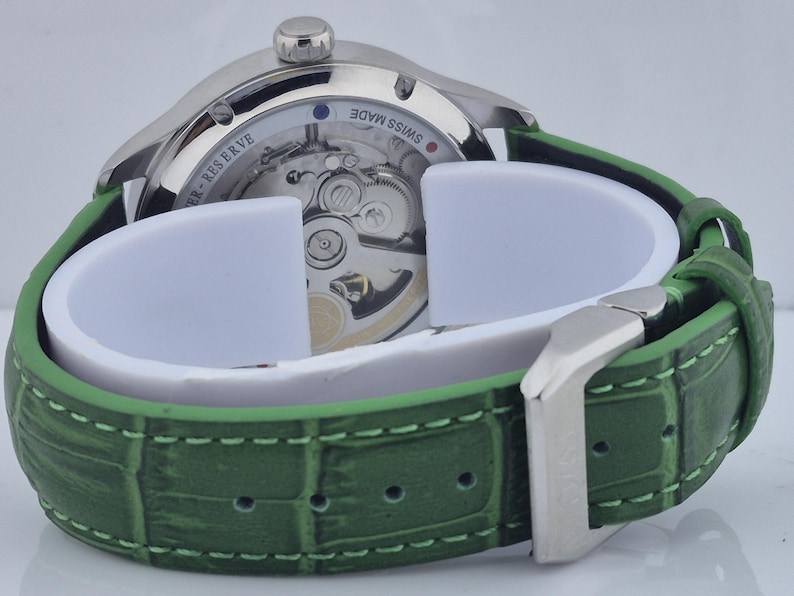 May include: A silver-toned wristwatch with a green leather band. The watch face is visible, showcasing the intricate internal mechanisms. The green band has a textured, crocodile-like pattern and a silver buckle.