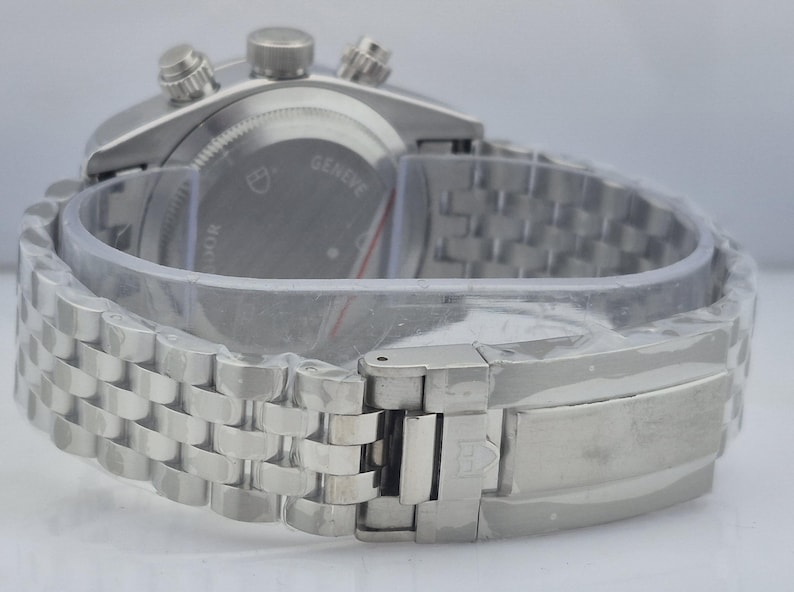 May include: A silver-toned wristwatch with a stainless steel band. The watch face is not visible, but the back of the watch is visible, with the word "Geneve" and other text. The watch is new and still has protective plastic.