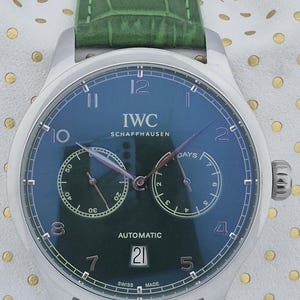 May include: A stainless steel IWC Schaffhausen automatic watch with a dark green face and a green leather strap. The watch face displays the date, day, and time. The watch has the text "IWC SCHAFFHAUSEN AUTOMATIC" on the face.