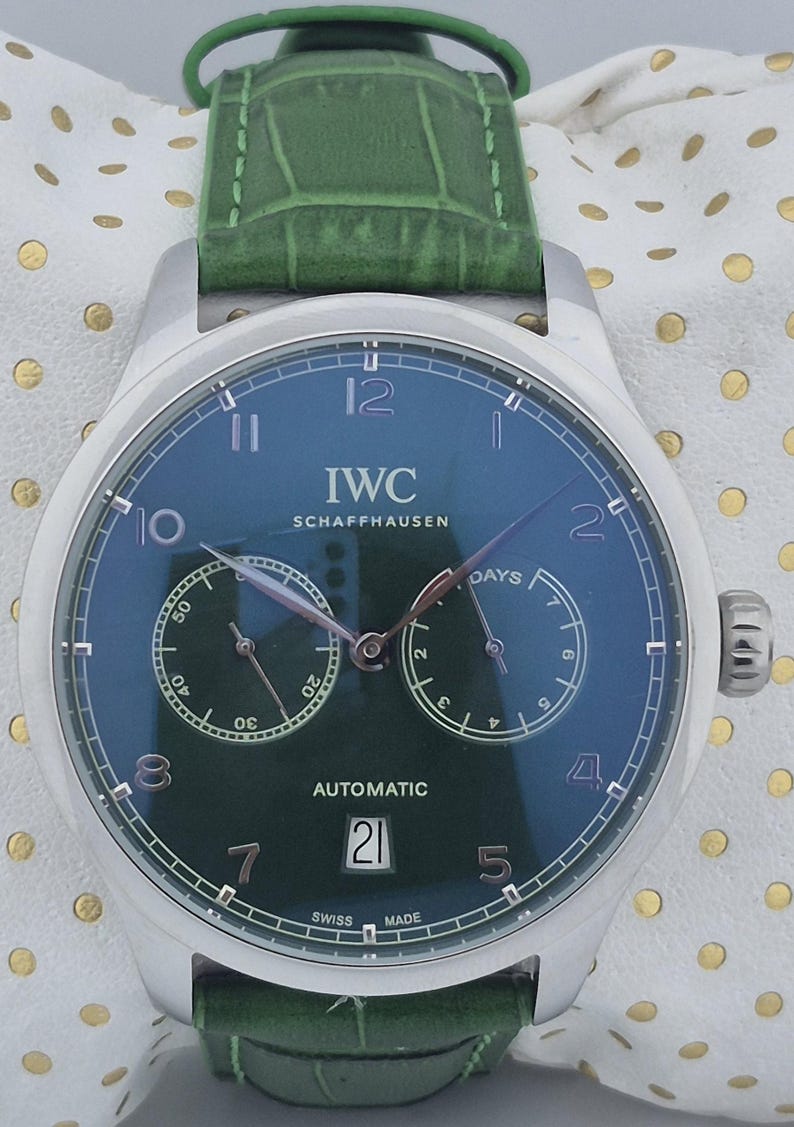 May include: A stainless steel IWC Schaffhausen automatic watch with a dark green face and a green leather strap. The watch face displays the date, day, and time. The watch has the text "IWC SCHAFFHAUSEN AUTOMATIC" on the face.