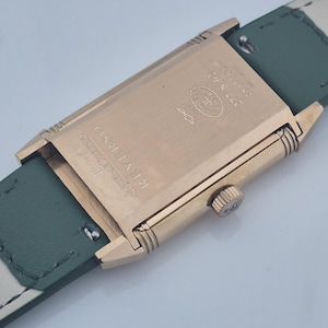 May include: Close-up of a gold-toned rectangular watch case with a green leather strap. The watch face is not visible. The back of the watch features the text "REVERSO" and other markings. The strap is approximately 0.75 inches wide.