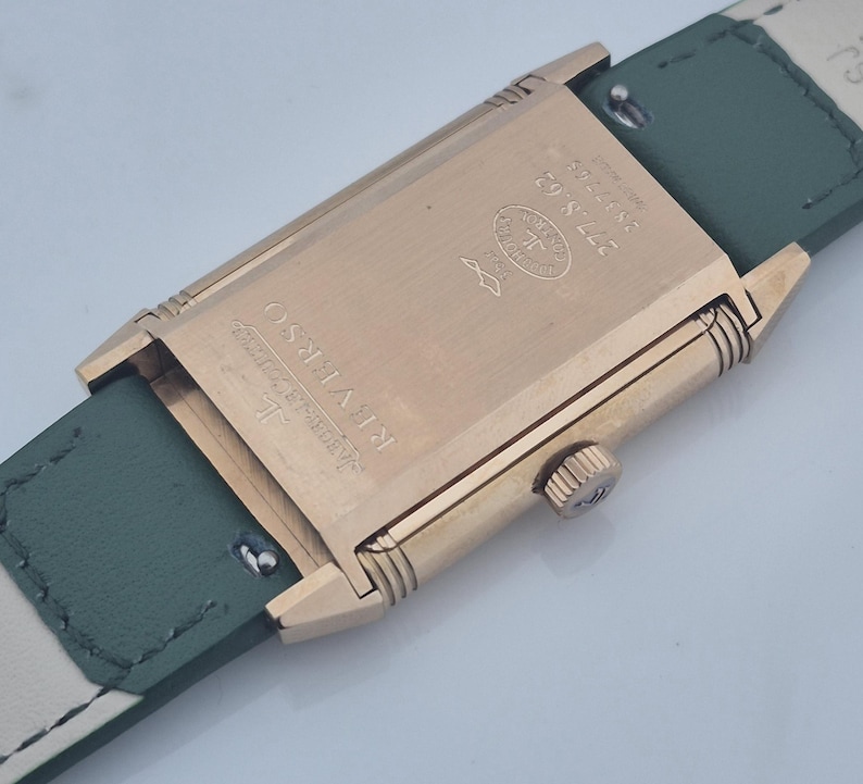 May include: Close-up of a gold-toned rectangular watch case with a green leather strap. The watch face is not visible. The back of the watch features the text "REVERSO" and other markings. The strap is approximately 0.75 inches wide.