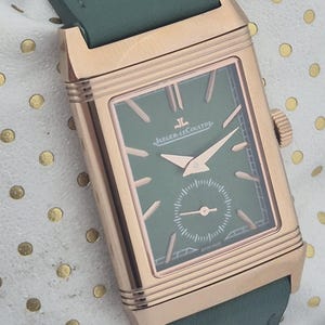 May include: A rectangular rose gold watch with a green face and hands, and a green leather strap. The watch face has the brand name "Jaeger-LeCoultre" printed on it. The watch is on a white surface with gold polka dots.