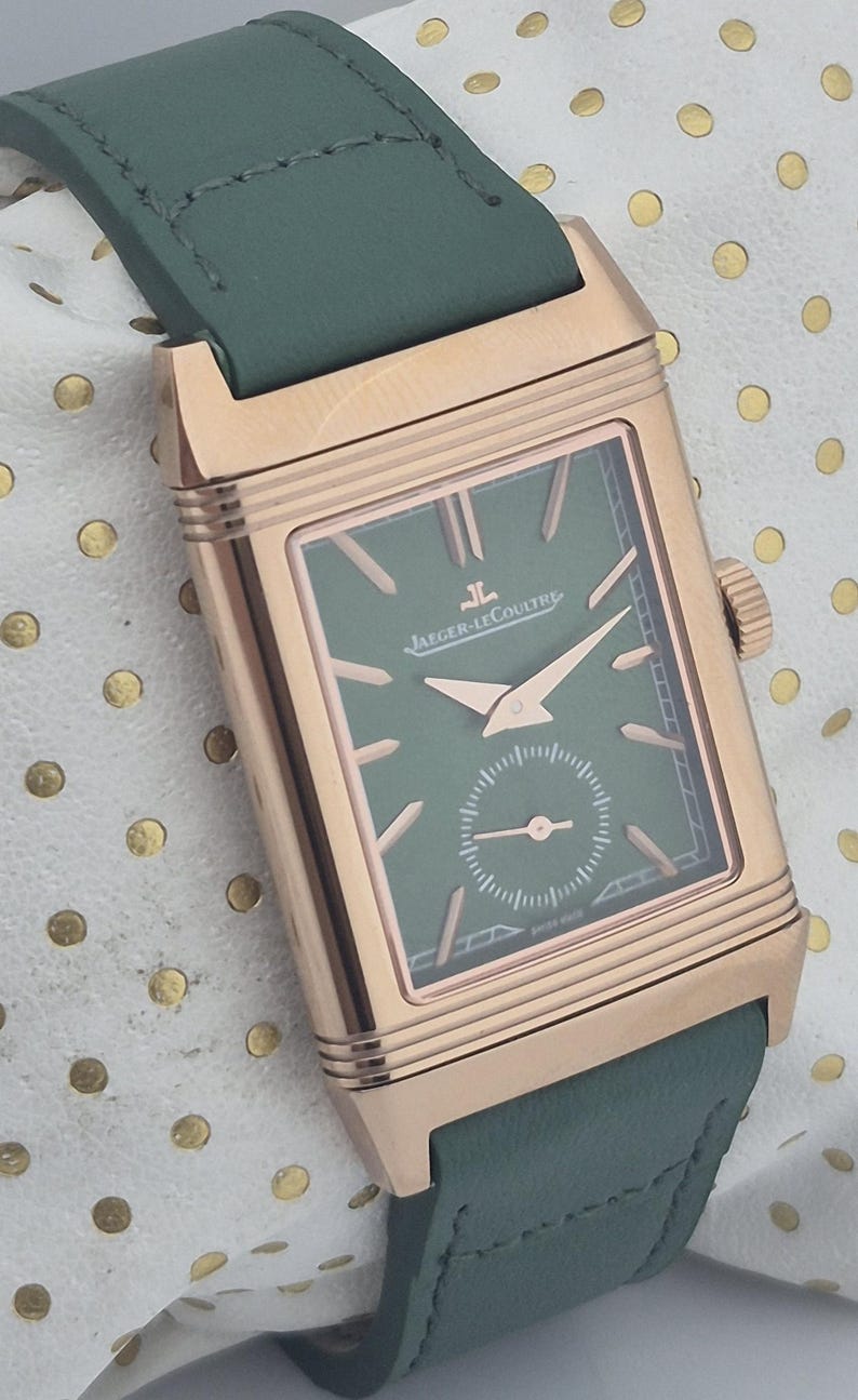 May include: A rectangular rose gold watch with a green face and hands, and a green leather strap. The watch face has the brand name "Jaeger-LeCoultre" printed on it. The watch is on a white surface with gold polka dots.