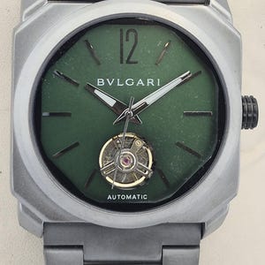 Może przedstawiać: Srebrny zegarek na rękę z kwadratową tarczą i zieloną tarczą. Zegarek posiada markę "BVLGARI" i napis "AUTOMATIC". Pasek zegarka wykonany jest z tego samego srebrnego metalu co koperta zegarka.