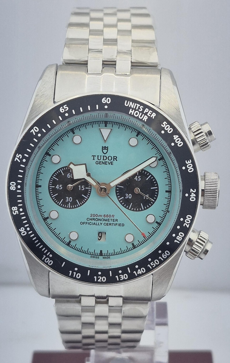 May include: A stainless steel wristwatch with a light blue face and black bezel. The watch features a chronograph function with two sub-dials and a date window. The watch band is made of stainless steel.