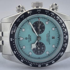 May include: A silver-toned wristwatch with a light blue face and a black bezel. The watch features three sub-dials, a date window, and a stainless steel band. The watch face displays the brand name "TUDOR GENEVE".