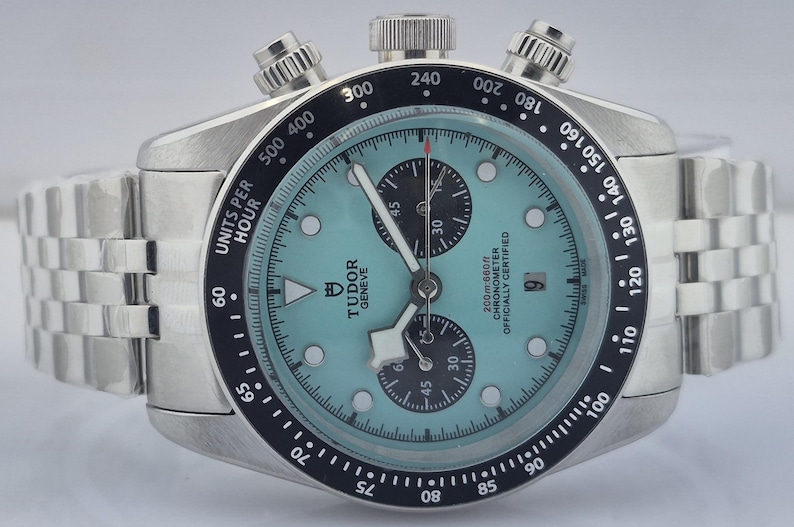 May include: A silver-toned wristwatch with a light blue face and a black bezel. The watch features three sub-dials, a date window, and a stainless steel band. The watch face displays the brand name "TUDOR GENEVE".