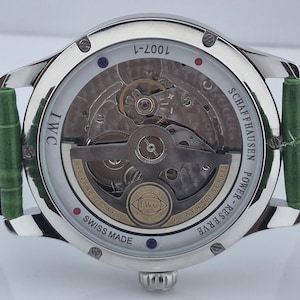 May include: A silver-toned IWC Schaffhausen watch with a transparent back, showcasing the intricate inner workings. The watch features a green leather strap and the text "SWISS MADE". The watch also has the text "POWER-RESERVE" and "SEVEN (7) DAYS".