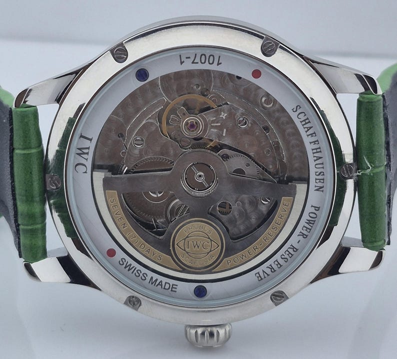 May include: A silver-toned IWC Schaffhausen watch with a transparent back, showcasing the intricate inner workings. The watch features a green leather strap and the text "SWISS MADE". The watch also has the text "POWER-RESERVE" and "SEVEN (7) DAYS".