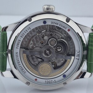 May include: Close-up of a silver-toned wristwatch with a transparent back, revealing intricate mechanical components. The watch features a green leather strap and the text "SWISS MADE" and "IWC". The watch face displays the text "SCHAFFHAUSEN POWER-RESERVE".