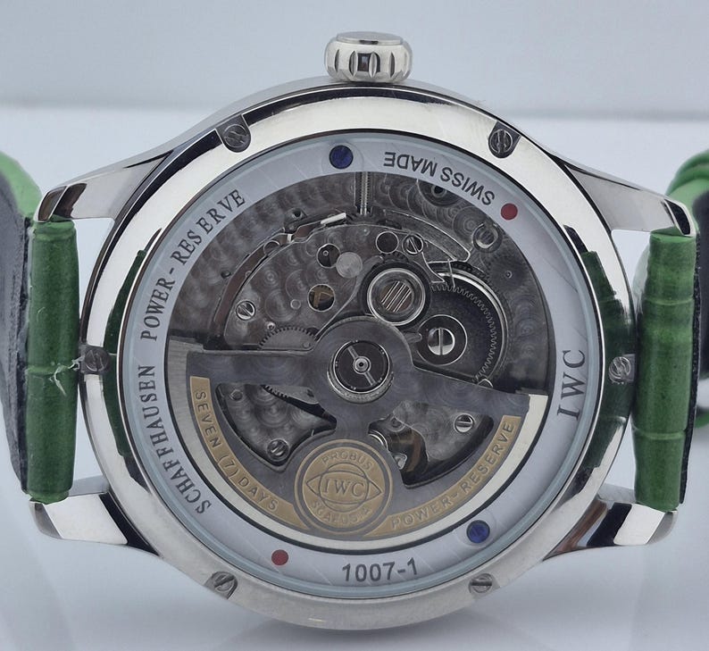 May include: Close-up of a silver-toned wristwatch with a transparent back, revealing intricate mechanical components. The watch features a green leather strap and the text "SWISS MADE" and "IWC". The watch face displays the text "SCHAFFHAUSEN POWER-RESERVE".