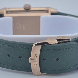 May include: Close-up of a luxury watch with a green leather strap and gold-colored buckle. The watch face is rectangular with a gold-colored case. The strap is secured with a gold-colored clasp.