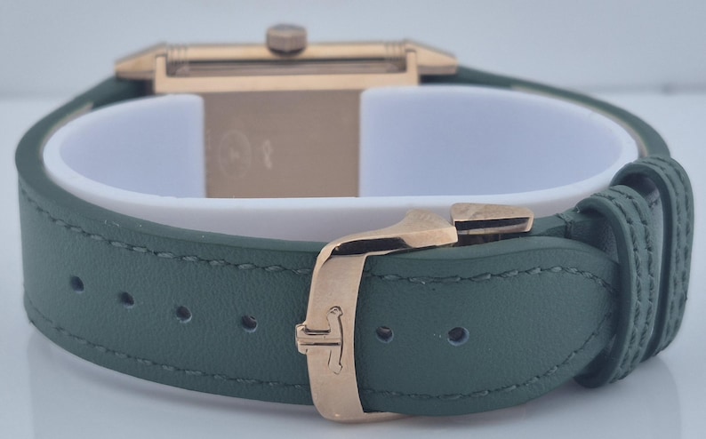 May include: Close-up of a luxury watch with a green leather strap and gold-colored buckle. The watch face is rectangular with a gold-colored case. The strap is secured with a gold-colored clasp.