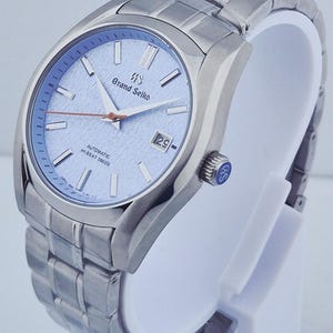 May include: A stainless steel Grand Seiko automatic wristwatch with a light blue dial. The watch features a date window at the 3 o'clock position and a silver-tone bracelet. The watch is displayed on a white stand.