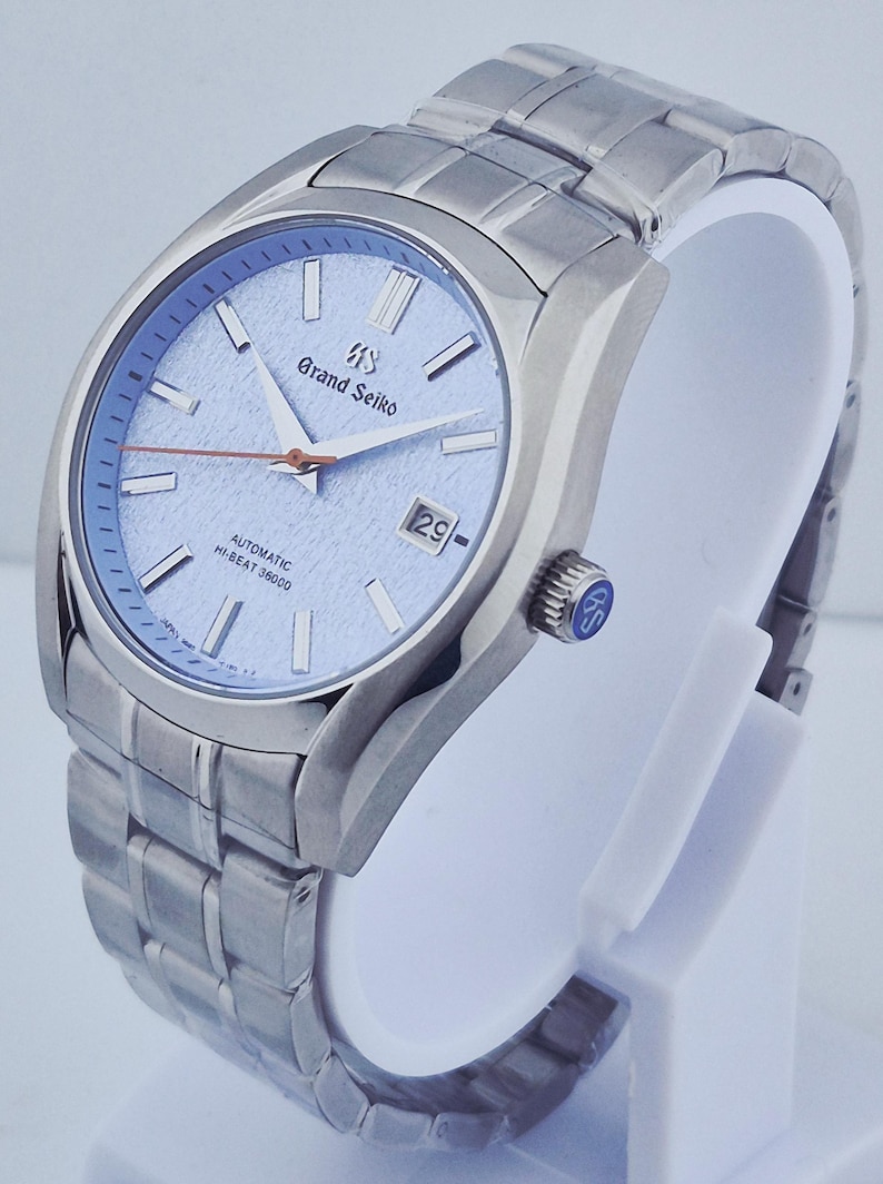 May include: A stainless steel Grand Seiko automatic wristwatch with a light blue dial. The watch features a date window at the 3 o'clock position and a silver-tone bracelet. The watch is displayed on a white stand.