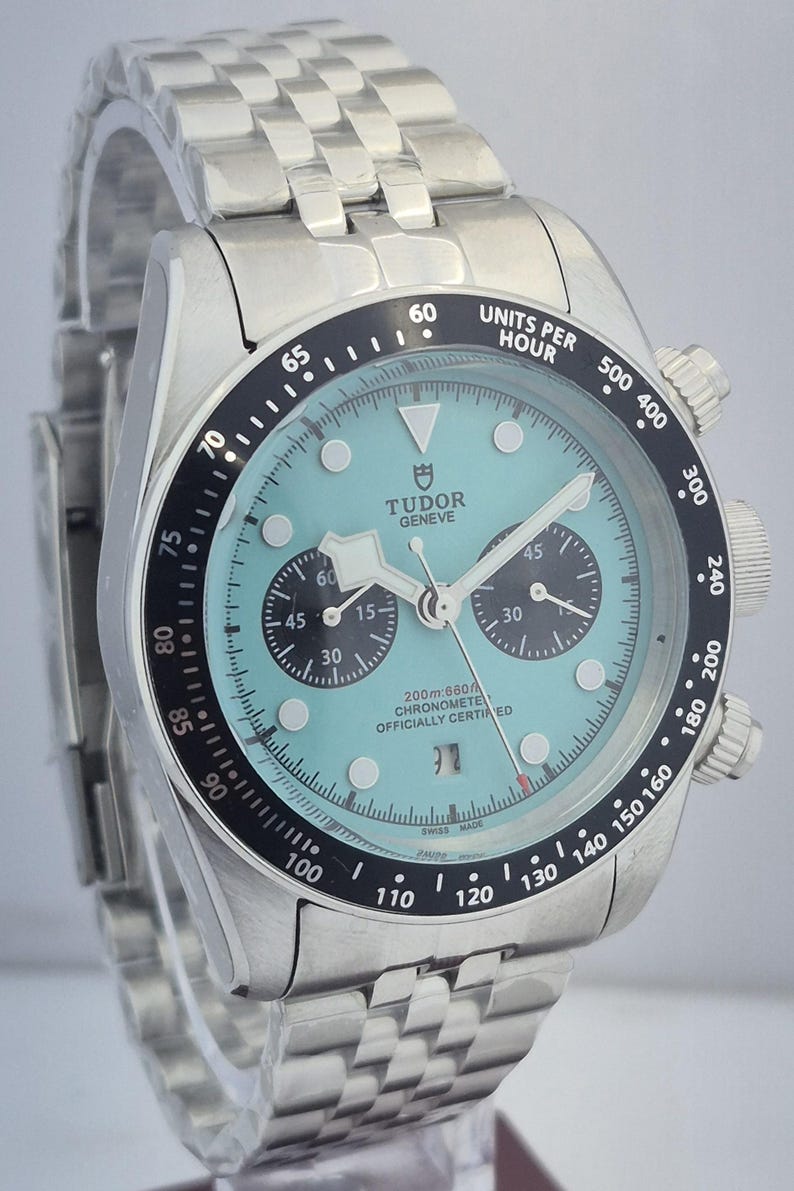 May include: A stainless steel Tudor Geneve chronograph watch with a light blue dial, black bezel, and a stainless steel bracelet. The watch features subdials, a date window, and is officially chronometer certified.