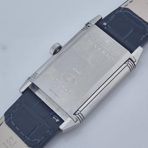 Pu&ograve; includere: Un orologio Jaeger-LeCoultre Reverso in acciaio inossidabile con cinturino in pelle blu scuro. Il quadrante rettangolare mostra "Jaeger-LeCoultre Reverso" e il numero di modello 277.S.62. Il cinturino &egrave; largo circa 2 cm.