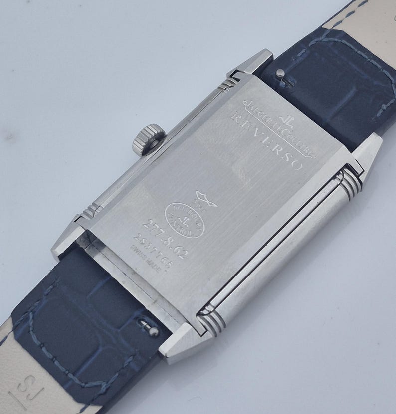 Pu&ograve; includere: Un orologio Jaeger-LeCoultre Reverso in acciaio inossidabile con cinturino in pelle blu scuro. Il quadrante rettangolare mostra "Jaeger-LeCoultre Reverso" e il numero di modello 277.S.62. Il cinturino &egrave; largo circa 2 cm.