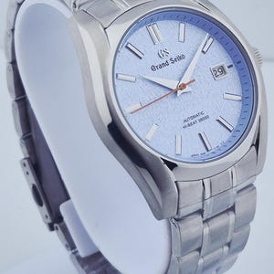 May include: A silver-toned wristwatch with a light blue dial. The watch features a date display at the 3 o'clock position and a silver-toned bracelet. The watch face has the words "Grand Seiko" and "Automatic Hi-Beat 36000".