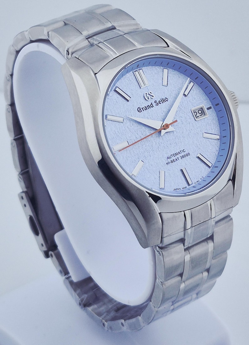 May include: A silver-toned wristwatch with a light blue dial. The watch features a date display at the 3 o'clock position and a silver-toned bracelet. The watch face has the words "Grand Seiko" and "Automatic Hi-Beat 36000".
