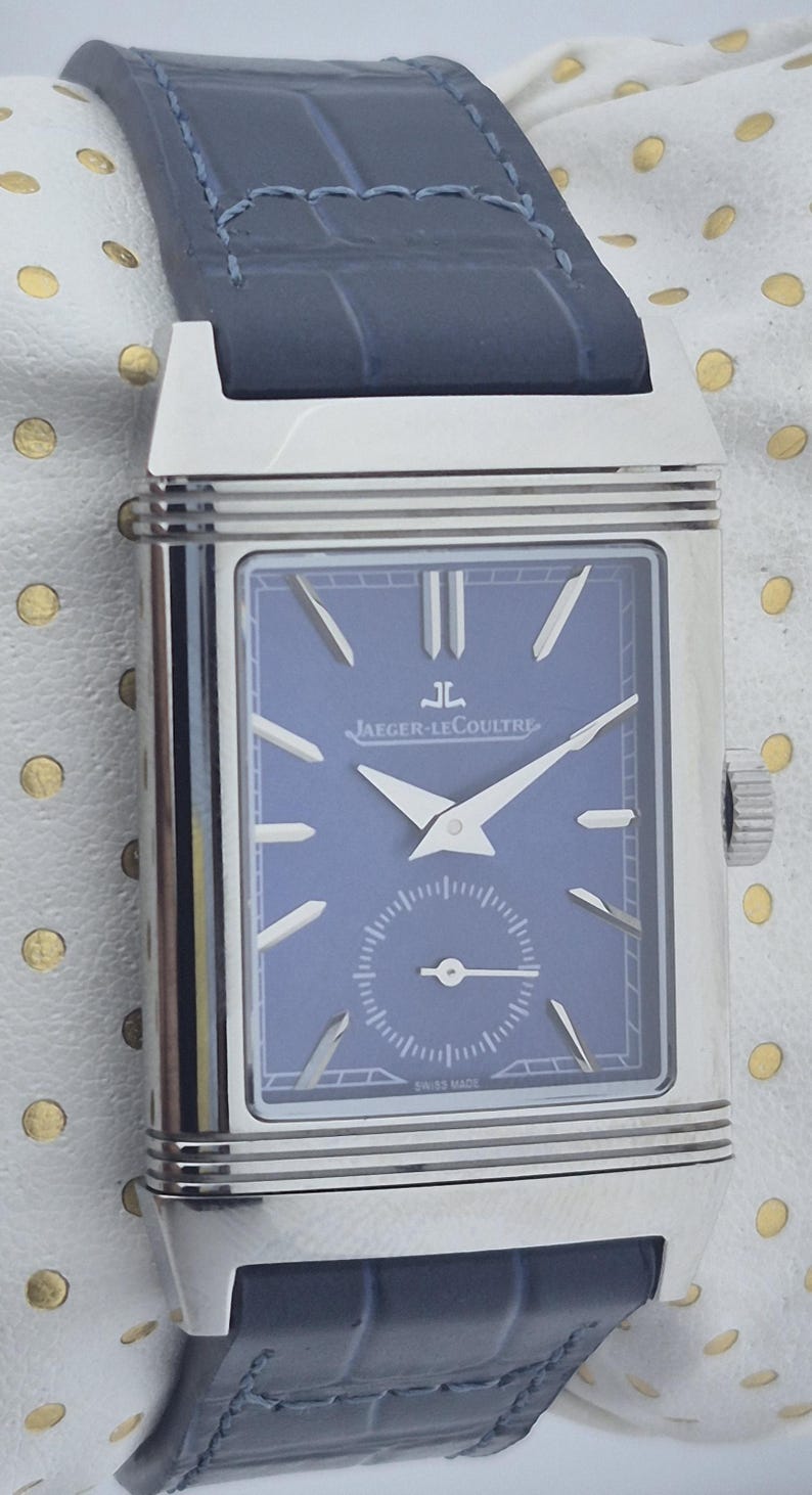 Pu&ograve; includere: Un orologio da polso rettangolare con quadrante blu e lancette argentate, con il logo Jaeger-LeCoultre. L'orologio ha una cassa argentata e un cinturino in pelle blu. Il quadrante include un piccolo quadrante dei secondi e la scritta "SWISS MADE".