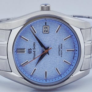 May include: A stainless steel Grand Seiko wristwatch with a light blue textured dial. The watch features silver-tone hour markers and hands, a date display at the 3 o'clock position, and a silver-tone bracelet. The watch face has the text "Grand Seiko" and "Automatic Hi-Beat 36000".