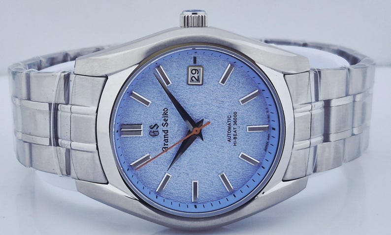 May include: A stainless steel Grand Seiko wristwatch with a light blue textured dial. The watch features silver-tone hour markers and hands, a date display at the 3 o'clock position, and a silver-tone bracelet. The watch face has the text "Grand Seiko" and "Automatic Hi-Beat 36000".