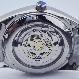May include: Close-up of a silver-toned wristwatch, showcasing the intricate inner workings through a transparent back. The watch features a detailed mechanism with gears and a decorative emblem. The crown is blue, and the watchband is made of metal.