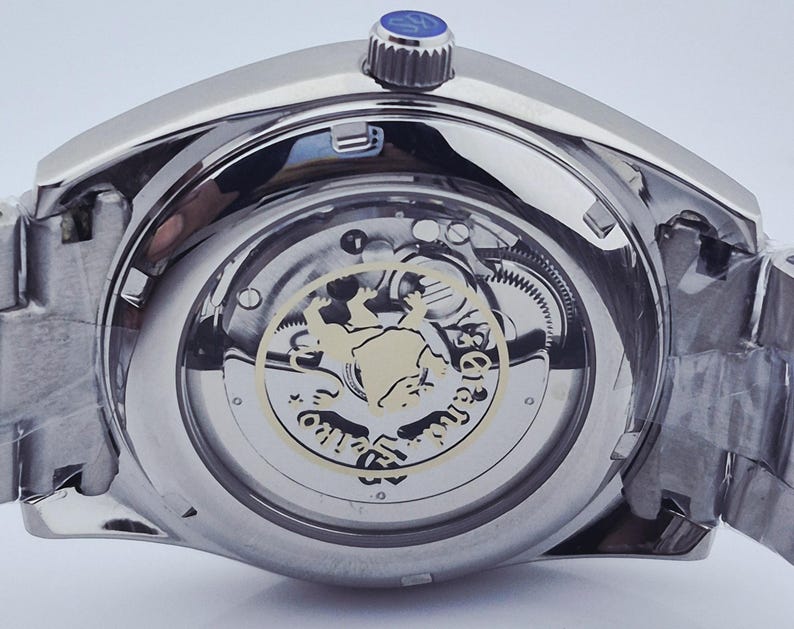 May include: Close-up of a silver-toned wristwatch, showcasing the intricate inner workings through a transparent back. The watch features a detailed mechanism with gears and a decorative emblem. The crown is blue, and the watchband is made of metal.