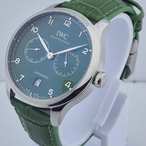 May include: A luxury wristwatch with a green face and a green leather band. The watch has a silver case and features the brand name "IWC SCHAFFHAUSEN". The watch face displays the date and two sub-dials. The band is approximately 8 inches long.