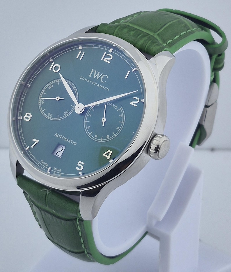 May include: A luxury wristwatch with a green face and a green leather band. The watch has a silver case and features the brand name "IWC SCHAFFHAUSEN". The watch face displays the date and two sub-dials. The band is approximately 8 inches long.