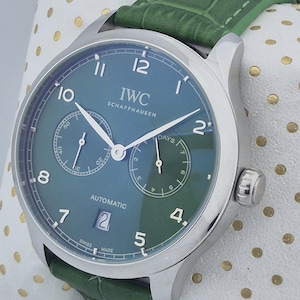 May include: A stainless steel IWC Schaffhausen automatic watch with a teal face and green leather strap. The watch face features two sub-dials, a date window, and the text "IWC SCHAFFHAUSEN". The watch is displayed on a white surface with gold polka dots.