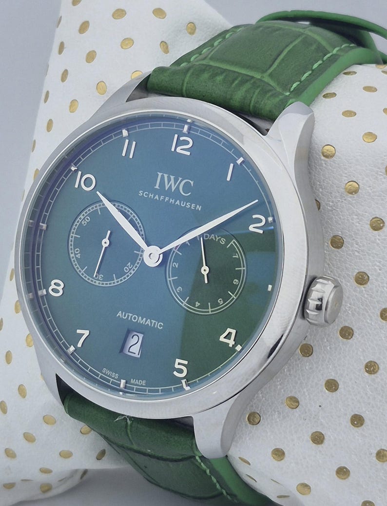 May include: A stainless steel IWC Schaffhausen automatic watch with a teal face and green leather strap. The watch face features two sub-dials, a date window, and the text "IWC SCHAFFHAUSEN". The watch is displayed on a white surface with gold polka dots.