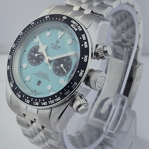 May include: A stainless steel Tudor Geneve watch with a light blue face, black bezel, and a stainless steel band. The watch features three sub-dials and a date display. The bezel has white markings for units per hour.