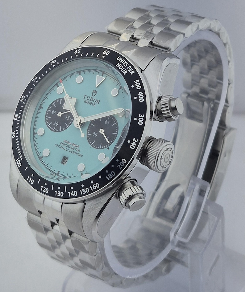 May include: A stainless steel Tudor Geneve watch with a light blue face, black bezel, and a stainless steel band. The watch features three sub-dials and a date display. The bezel has white markings for units per hour.