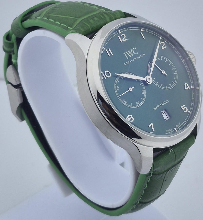 May include: A luxury wristwatch with a green dial and a green leather band. The watch has a silver-colored case and features the text "IWC SCHAFFHAUSEN" on the dial. The watch also has two sub-dials and a date display.