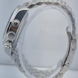 May include: A silver wristwatch with a stainless steel band. The watch face is black, and the watch has multiple buttons and dials. The band is made of small, interconnected links. The watch is displayed on a clear, curved stand.