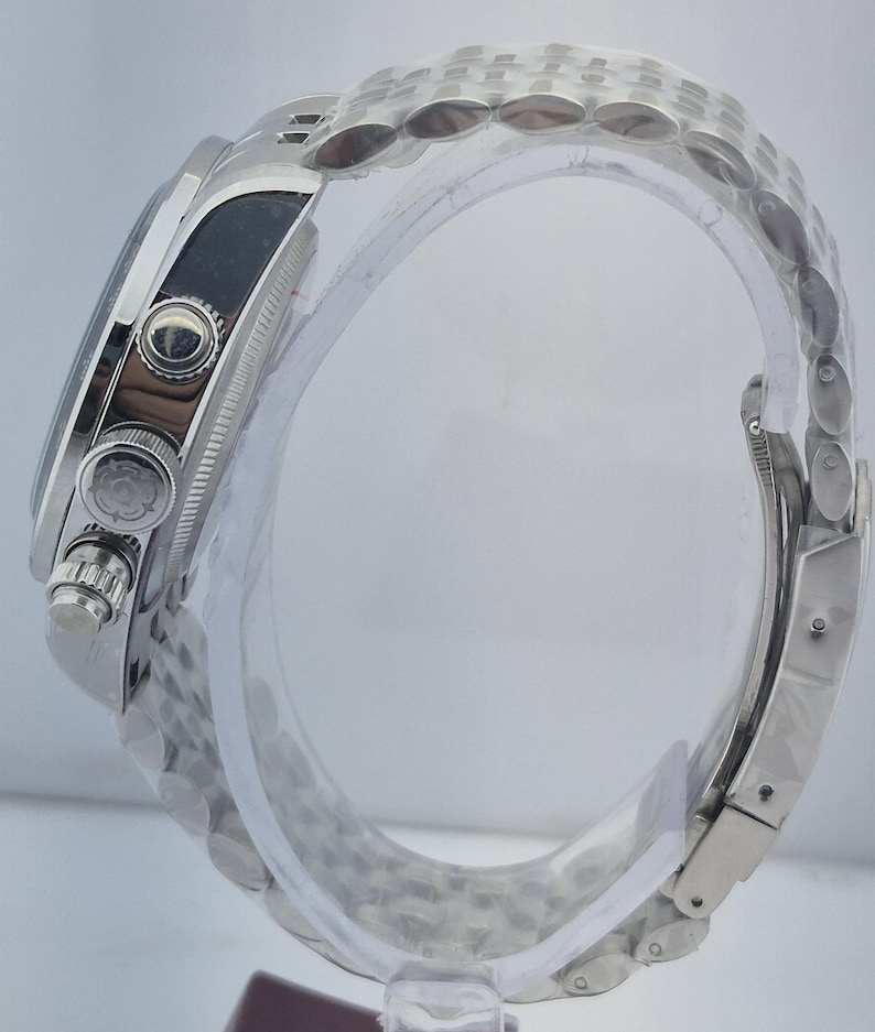 May include: A silver wristwatch with a stainless steel band. The watch face is black, and the watch has multiple buttons and dials. The band is made of small, interconnected links. The watch is displayed on a clear, curved stand.