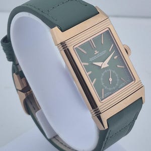May include: A rectangular wristwatch with a green face and a green leather band. The watch has a gold-colored case and a small seconds dial. The watch is displayed on a white stand.